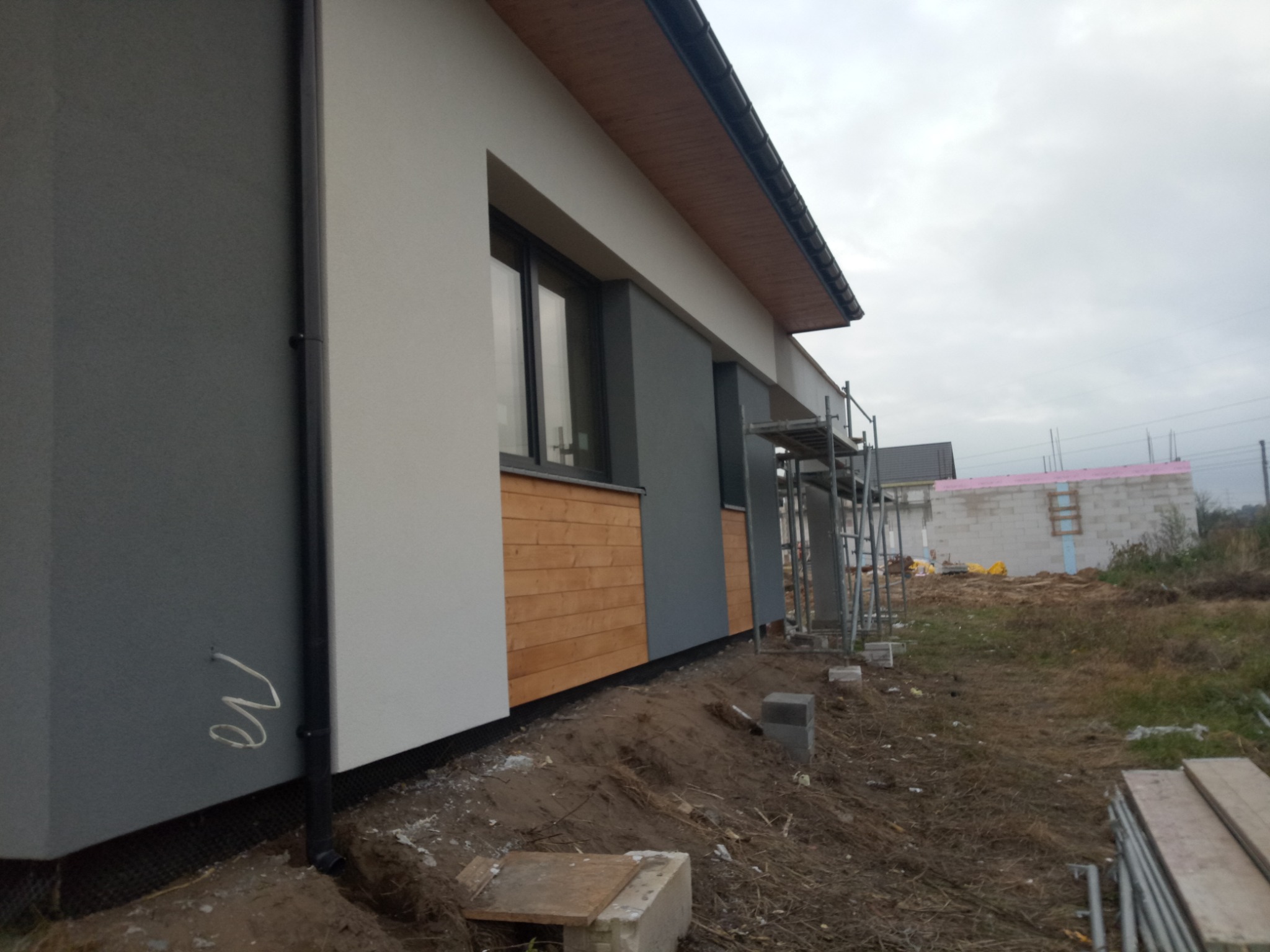 Fragment elewacji domu jednorodzinnego w trakcie wykańczania, z widocznym połączeniem tynku w odcieniach szarości i bieli oraz drewnianej okładziny, rusztowania i materiały budowlane wokół.