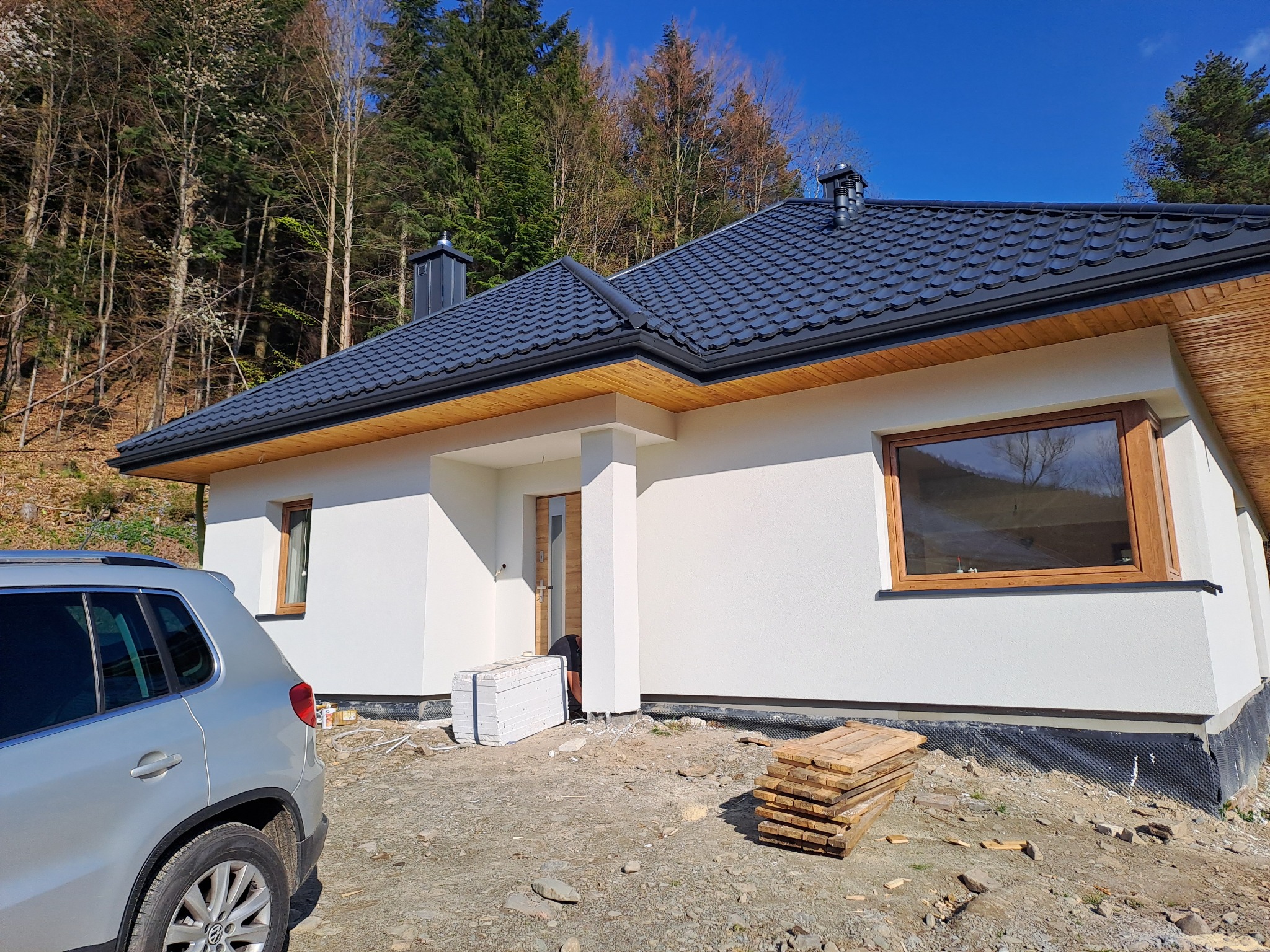 Biały dom jednorodzinny z ciemną dachówką i drewnianą podbitką, widok z zewnątrz, z częściowo wykonanymi pracami wykończeniowymi wokół budynku, w tle las.