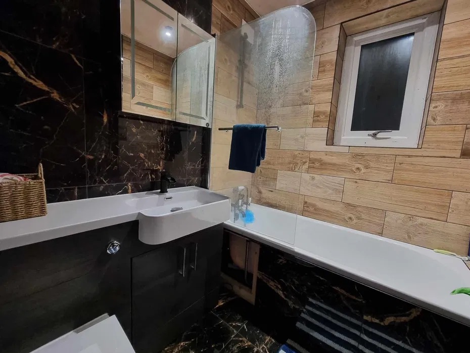 Wykończona łazienka z czarnymi marmurowymi płytkami, drewnopodobną okładziną ścienną, białą wanną i umywalką z szafką oraz oknem w nietypowej ramie.