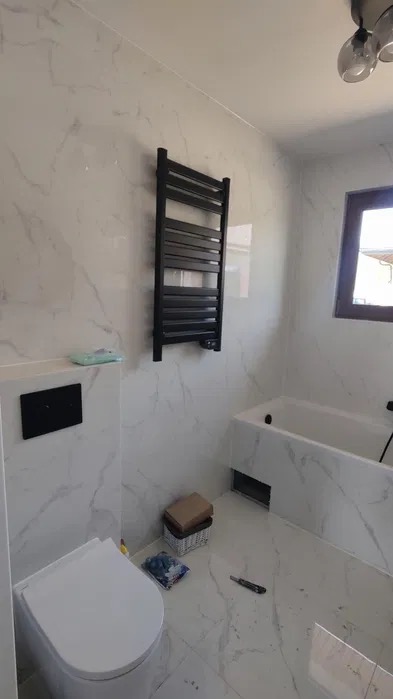 Wykończona łazienka z marmurowymi płytkami, czarnym grzejnikiem drabinkowym, wiszącą toaletą i zabudowaną wanną. Na podłodze leży nóż do tapet.