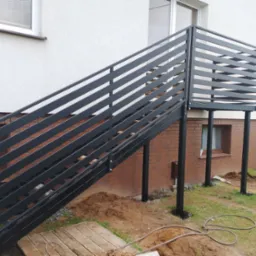 Zewnętrzne schody z balustradą z poziomych, czarnych profili, prowadzące do wejścia na podwyższeniu, z widocznym fragmentem elewacji budynku z cegły i białego tynku.