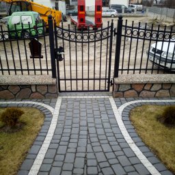 Stalmix - Brama wejściowa z czarnego metalu z ozdobnymi elementami, osadzona na kamiennych słupkach, prowadząca do posesji z szarej kostki brukowej z białymi obrzeżami, w tle budynki mieszkalne i sprzęt...