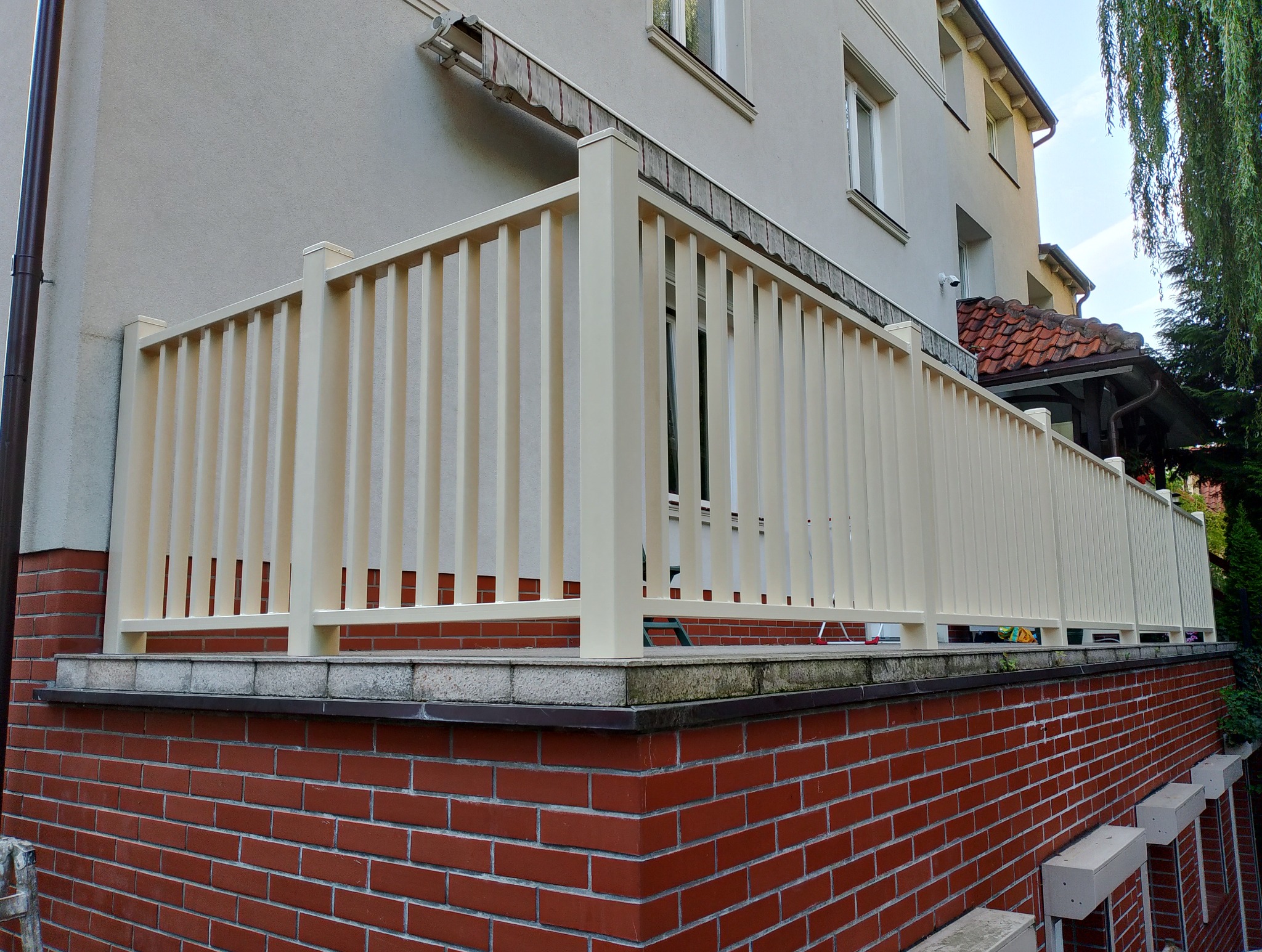 Beżowa balustrada z pionowych profili zamontowana na murku z czerwonej cegły, widok na dom z jasną elewacją i czerwonym dachem.