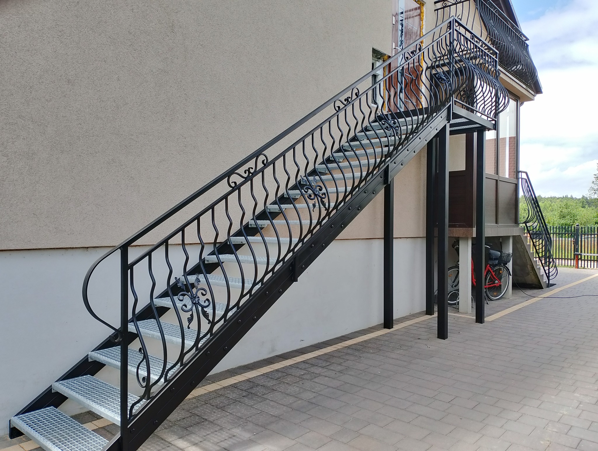 Zewnętrzne schody z czarną, kutą balustradą prowadzące na balkon, widoczne z boku, z rowerem zaparkowanym pod spodem.
