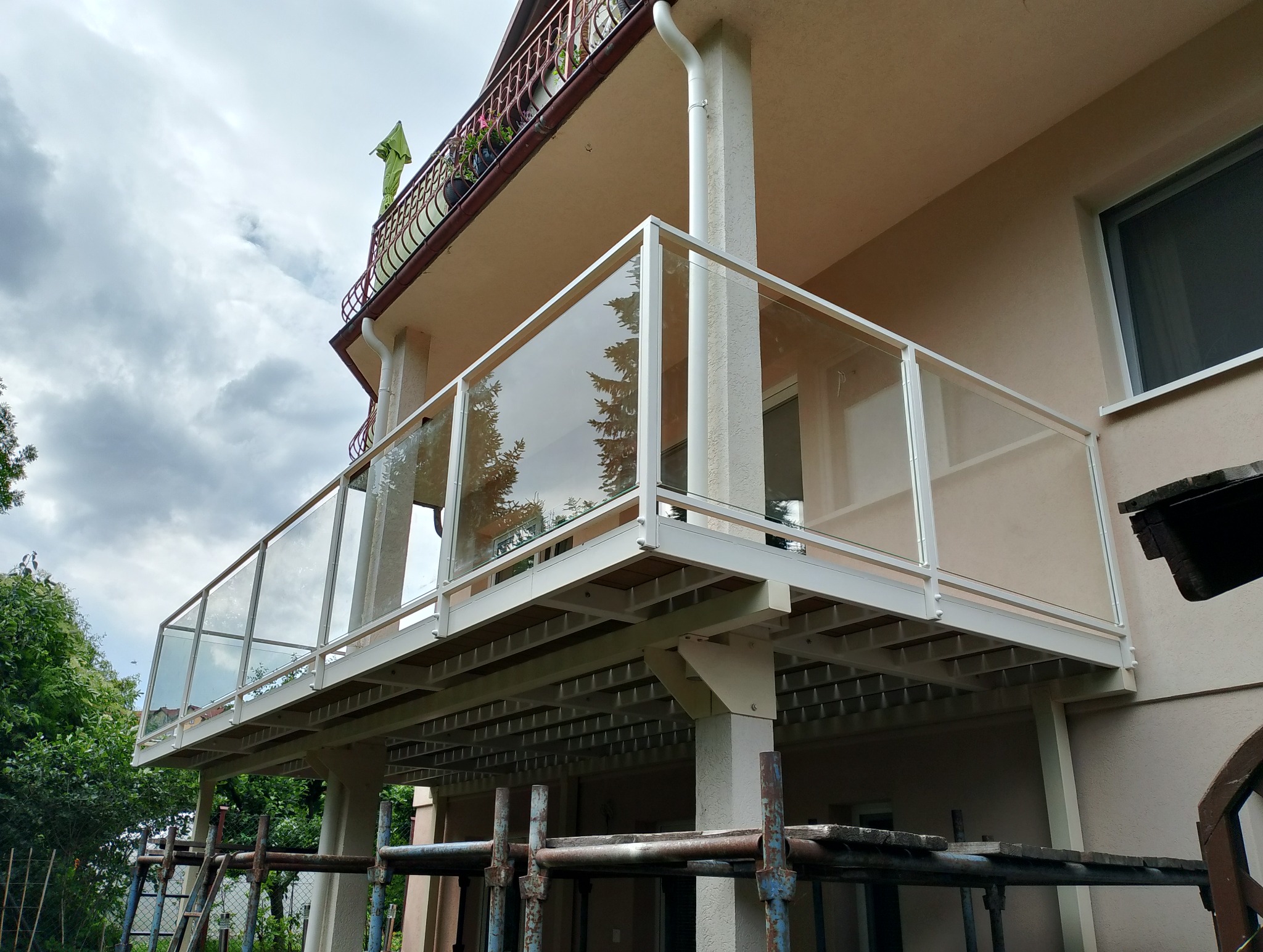 Balkon z nowoczesną, szklaną balustradą na metalowej konstrukcji, widoczny z dołu, z rusztowaniem w tle sugerującym prace wykończeniowe.