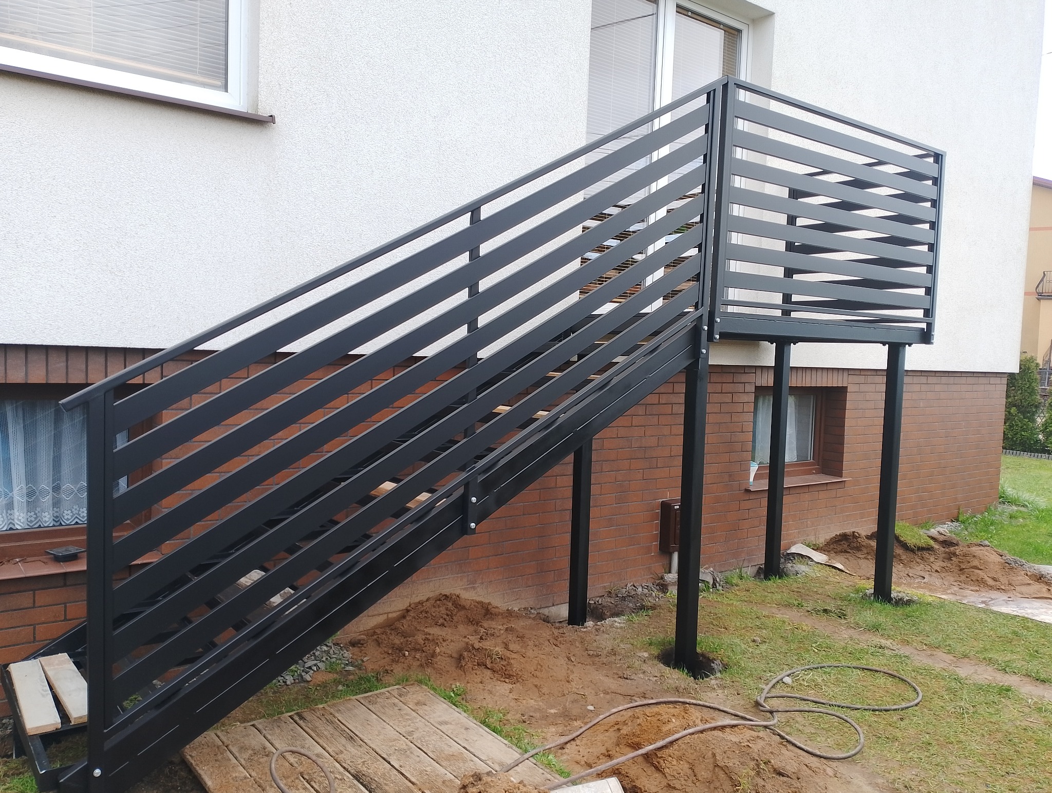 Zewnętrzne schody z balustradą z poziomych, czarnych profili, prowadzące do wejścia na podwyższeniu, z widocznym fragmentem elewacji budynku z cegły i białego tynku.