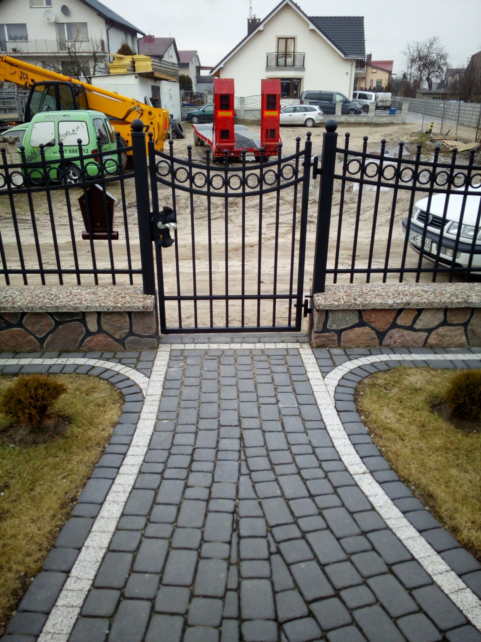 Brama wejściowa z czarnego metalu z ozdobnymi elementami, osadzona na kamiennych słupkach, prowadząca do posesji z szarej kostki brukowej z białymi obrzeżami, w tle budynki mieszkalne i sprzęt...