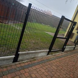 ŚLĄSKIE CENTRUM OGRODZEŃ TOMASZ BUCKI - Czarna brama panelowa z furtką, zamontowana na betonowej podmurówce, widoczna z perspektywy brukowanej ścieżki. W tle trawnik i budynek.