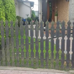 ŚLĄSKIE CENTRUM OGRODZEŃ TOMASZ BUCKI - Ciemne, drewniane ogrodzenie z dekoracyjnymi wycięciami, wsparte na ceglanych słupkach z daszkami, na tle zieleni i domu z cegły. Widok z zewnątrz posesji.