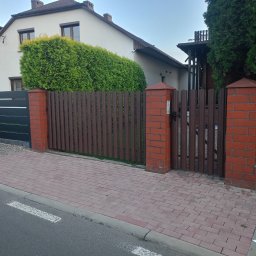 ŚLĄSKIE CENTRUM OGRODZEŃ TOMASZ BUCKI - Nowoczesne ogrodzenie posesji z brązowych paneli i ceglanych słupków, furtka z tego samego materiału. Częściowo widoczny szary samochód i chodnik z kostki.