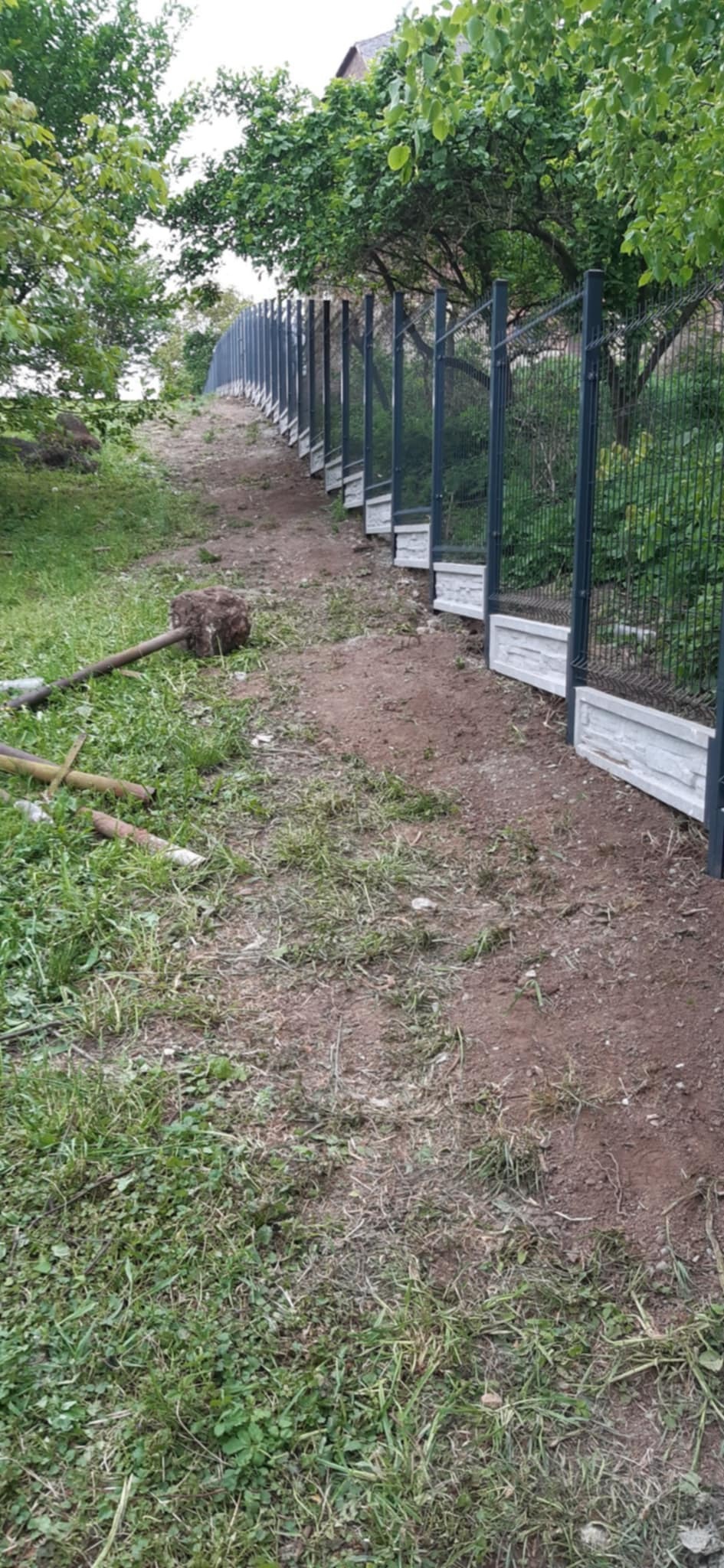 Metalowe panele ogrodzeniowe z podmurówką na nierównym terenie, widoczne narzędzia pracy. Świeżo zamontowane ogrodzenie na wzniesieniu.