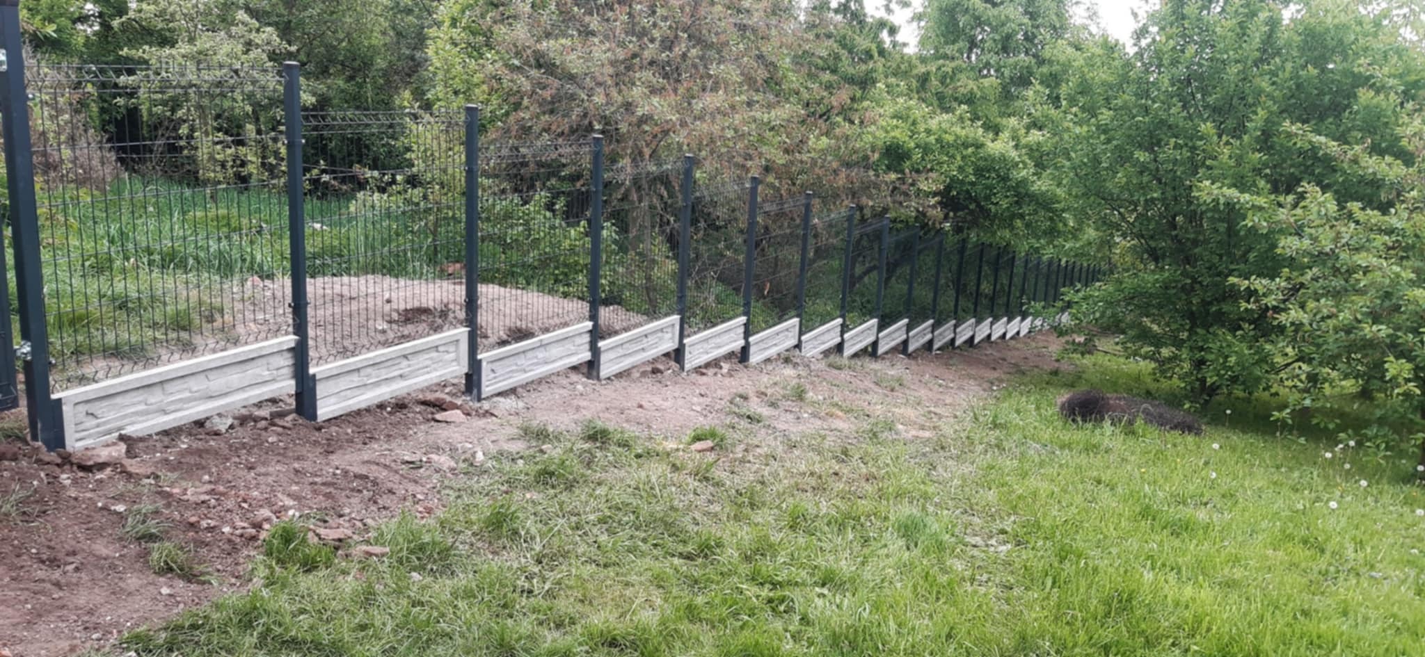 Długi, szary panelowy płot z betonową podmurówką, biegnący wzdłuż zielonego wzgórza, z drzewami w tle. Widok z dołu, ukazujący perspektywę i głębię.