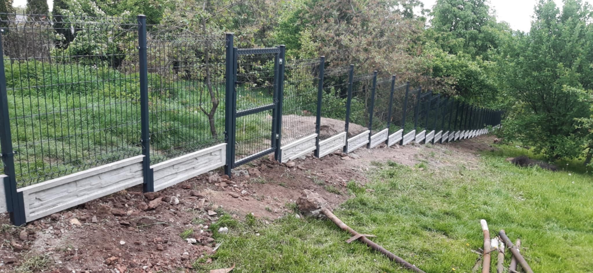 Panelowe ogrodzenie z bramką na nierównym terenie, z betonową podmurówką o fakturze kamienia. Widok z boku, w tle zieleń drzew.