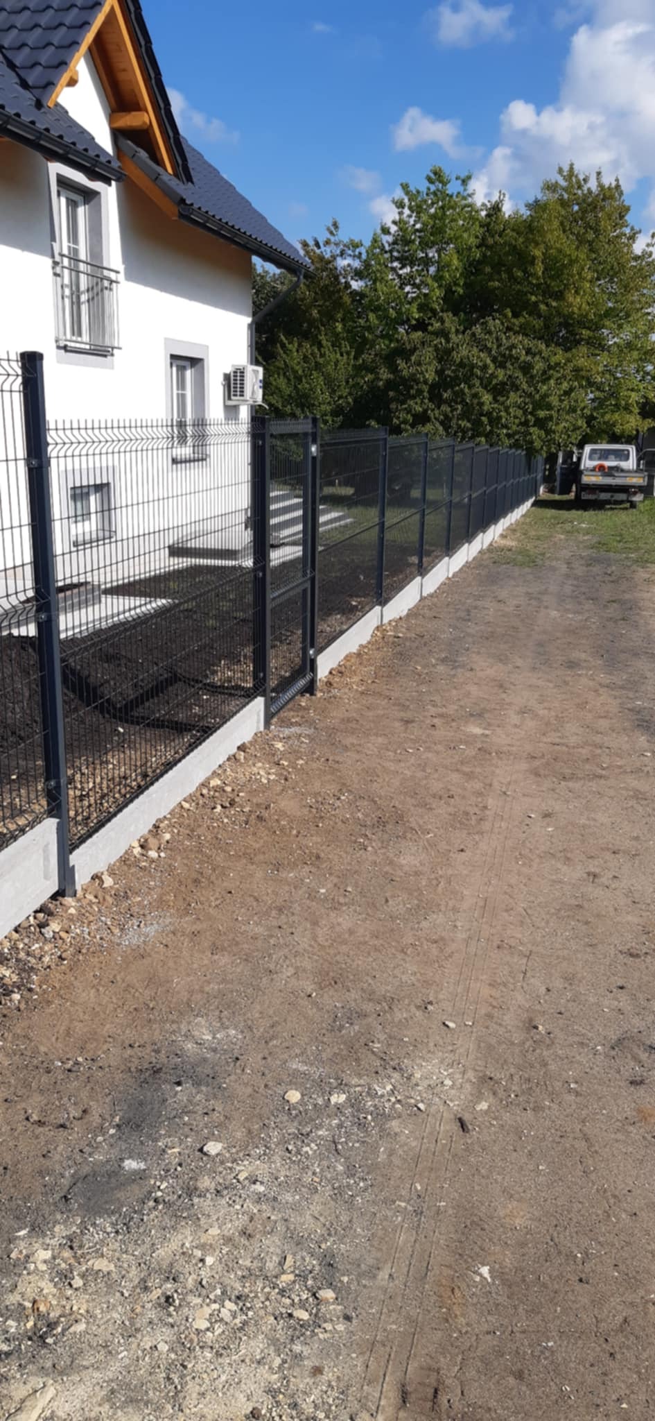 Nowoczesne ogrodzenie panelowe z bramką, zamontowane na betonowej podmurówce, otaczające posesję z białym domem i zielenią w tle. Słoneczny dzień.