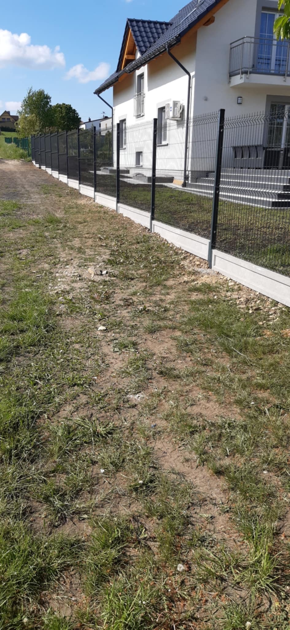 Nowoczesne, czarne ogrodzenie panelowe na betonowej podmurówce, otaczające biały dom z ciemnym dachem. Widok z perspektywy gruntu, podkreślający długość i solidność konstrukcji.