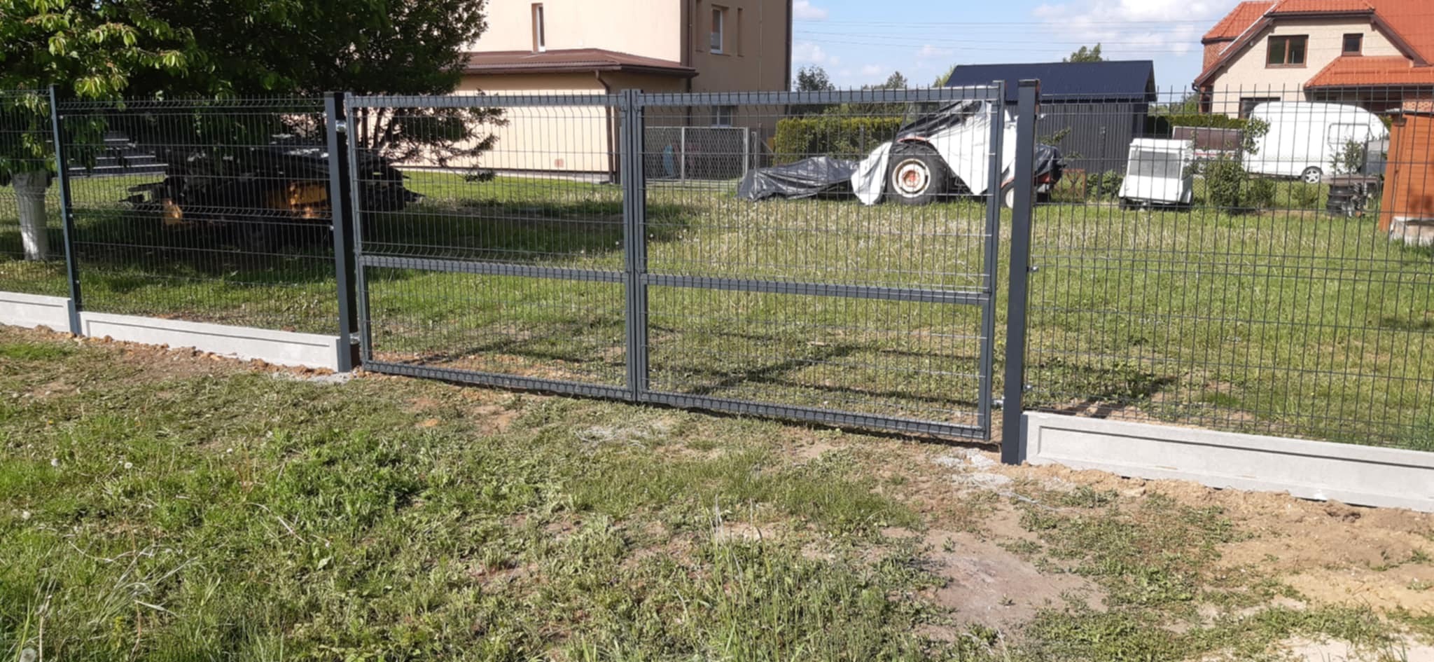 Szara, metalowa brama panelowa z betonową podmurówką na tle zielonej trawy i zabudowań mieszkalnych. Widoczne słupki i siatka ogrodzeniowa.