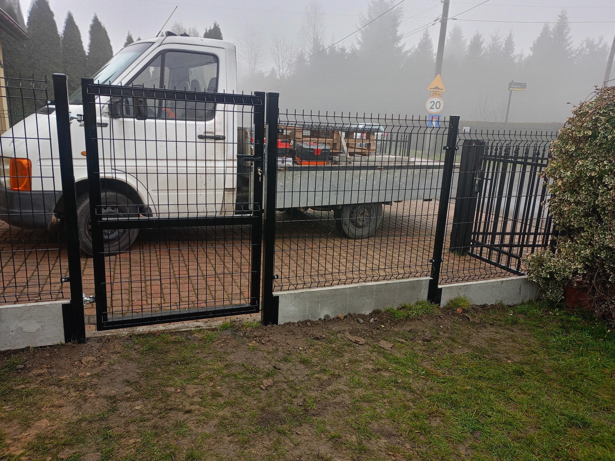 Nowoczesne, czarne ogrodzenie panelowe z furtką na betonowej podmurówce. W tle biały samochód dostawczy i zarośla w pochmurny dzień.