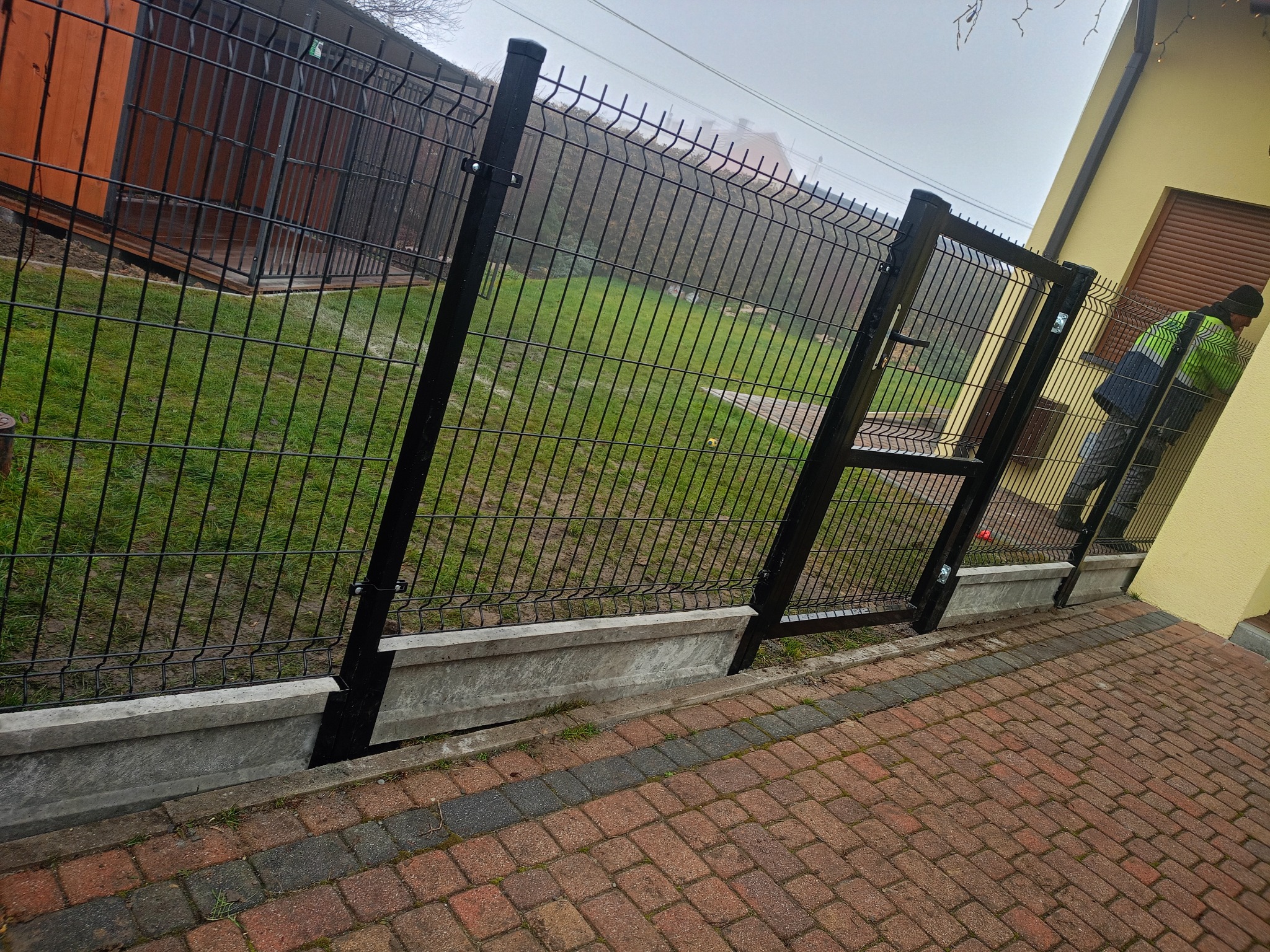 Czarna brama panelowa z furtką, zamontowana na betonowej podmurówce, widoczna z perspektywy brukowanej ścieżki. W tle trawnik i budynek.