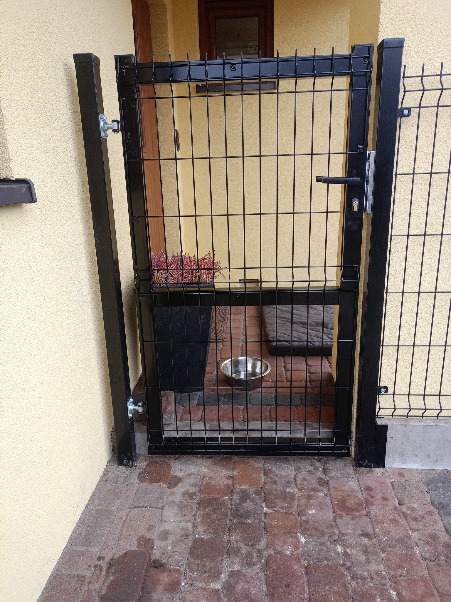 Czarna furtka z paneli ogrodzeniowych, zamontowana przy wejściu do domu. W tle widoczny kwiat w doniczce i miska dla zwierząt, co sugeruje przeznaczenie dla psa.