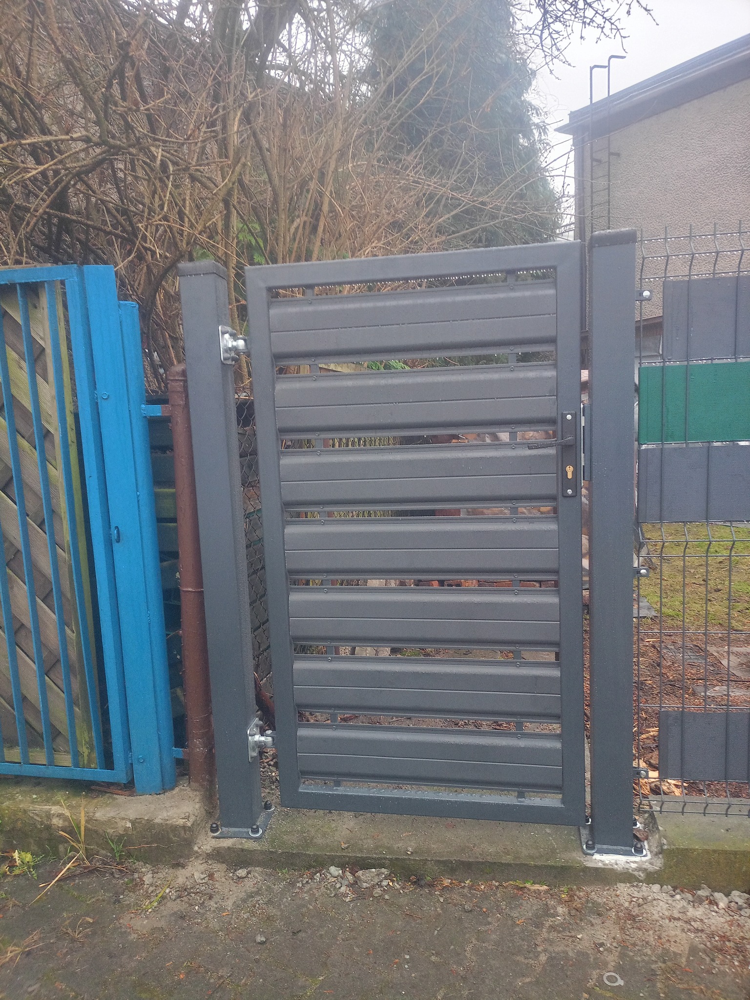 Nowoczesna, szara furtka z poziomych profili, zamontowana przy ogrodzeniu panelowym. Widoczne detale mocowania i zamek. Tło stanowi zieleń i zabudowa.