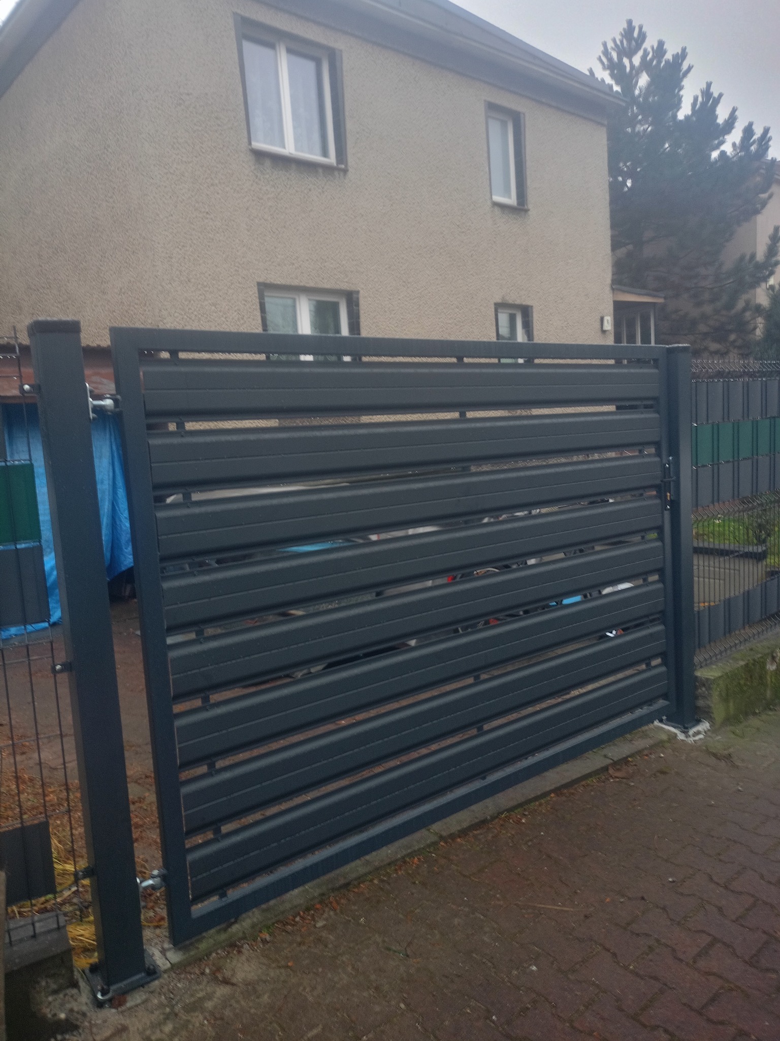 Nowoczesna, pozioma brama metalowa w kolorze grafitowym, zamontowana przy ogrodzeniu panelowym. Widoczny fragment domu w tle. Brama zamyka posesję.
