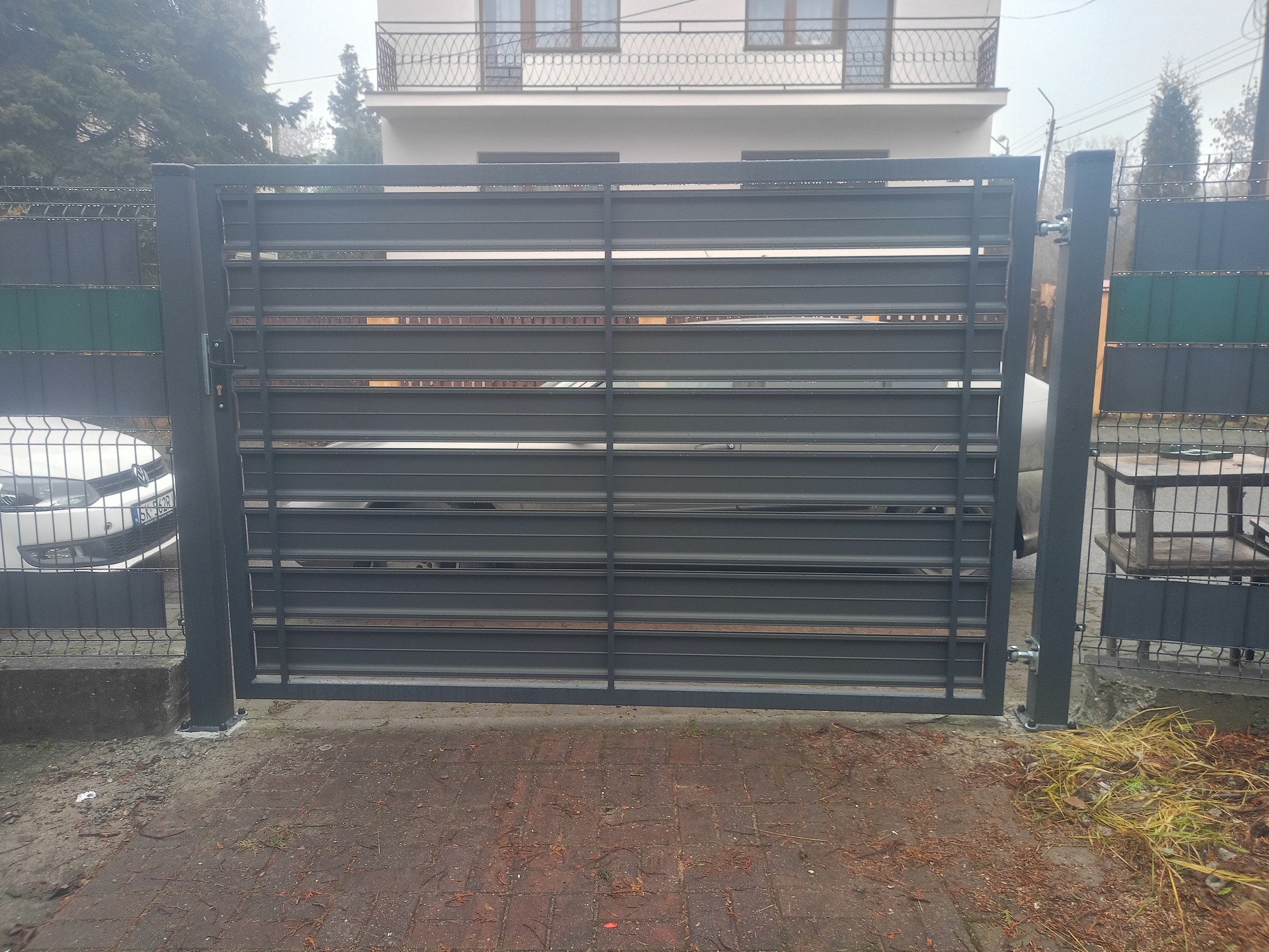 Nowoczesna, pozioma brama przesuwna z metalu w kolorze grafitowym, zamontowana przy ogrodzeniu panelowym. Widoczny fragment samochodu i dwupiętrowy dom w tle.