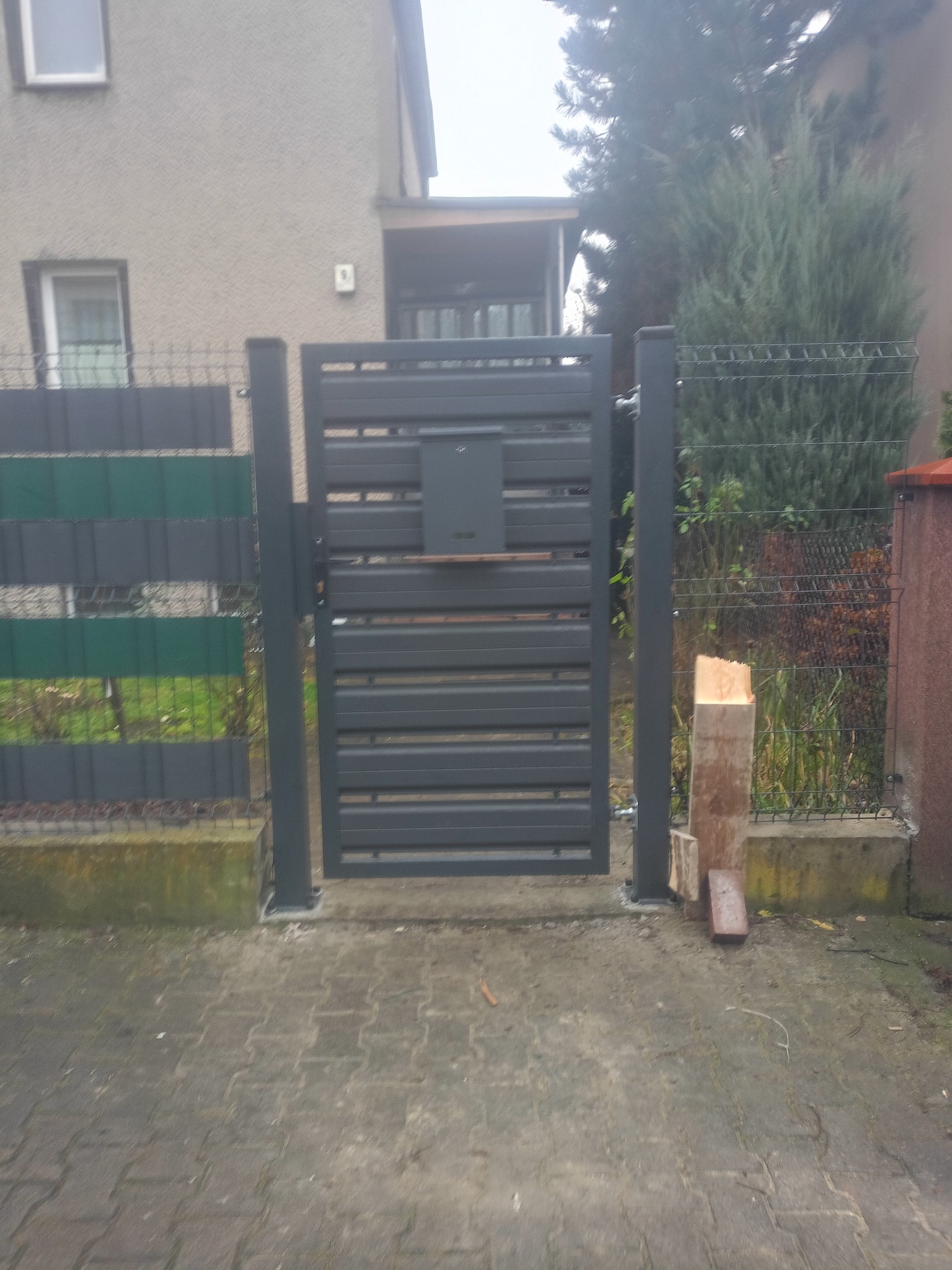 Nowoczesna, grafitowa furtka z poziomych profili, z zamontowaną skrzynką na listy, wkomponowana w ogrodzenie panelowe z elementami dekoracyjnymi.