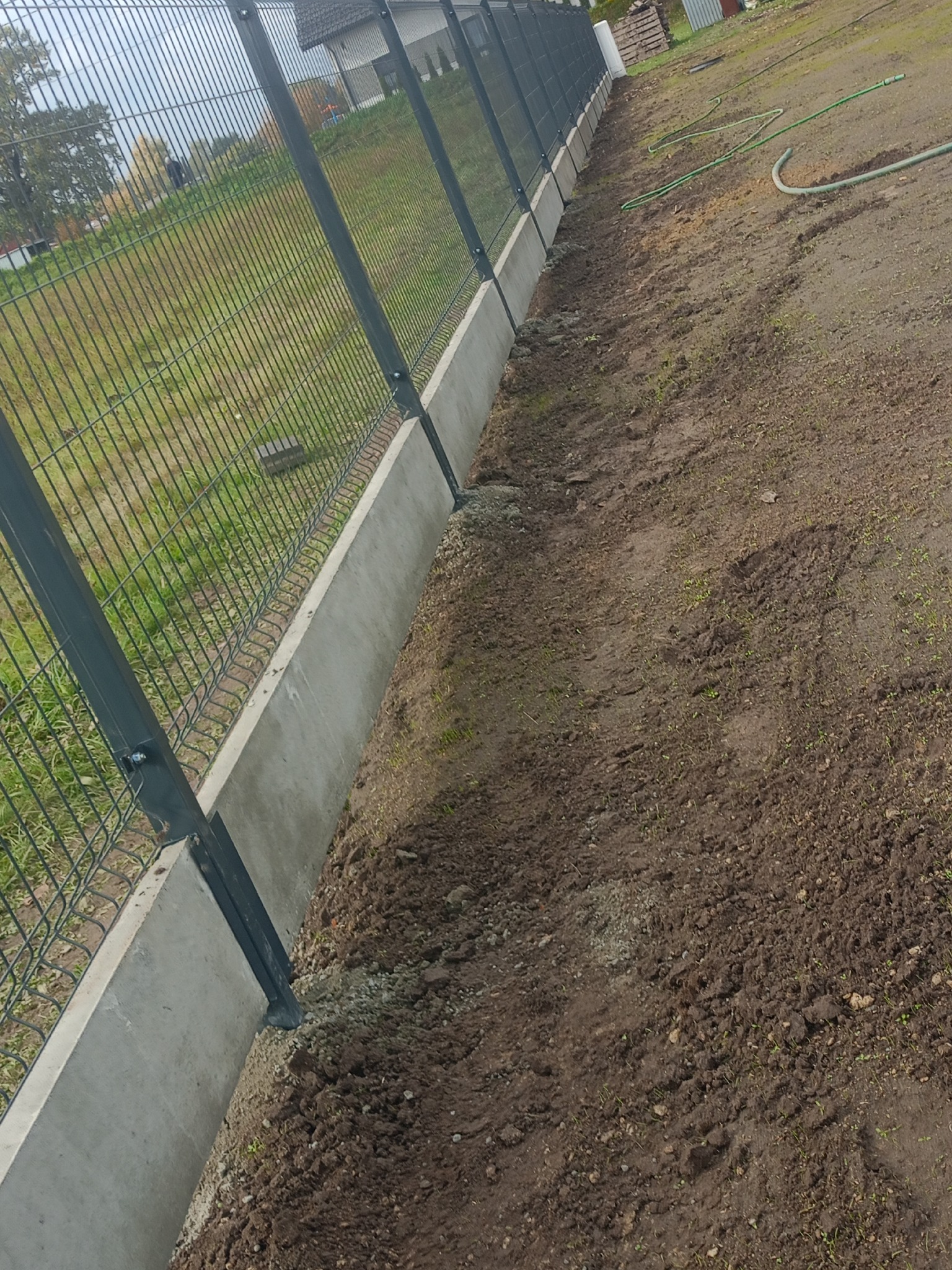 Nowoczesne ogrodzenie panelowe z podmurówką betonową, widok z bliska na świeżo obsypaną ziemią wzdłuż ogrodzenia. W tle dom i zieleń.