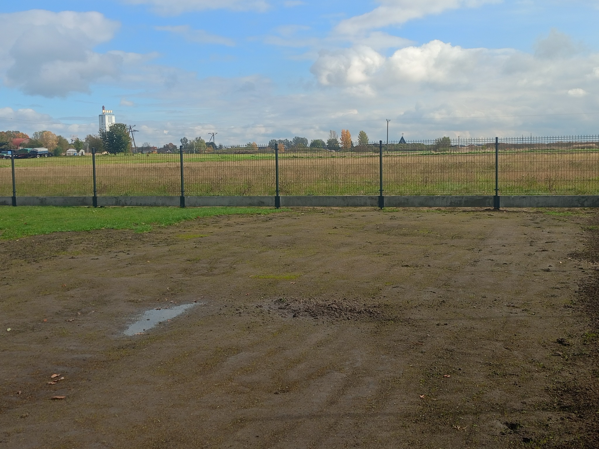 Nowoczesne ogrodzenie panelowe w kolorze grafitowym, oddzielające posesję od łąki. W tle widoczny budynek z napisem PIAST i błękitne niebo z chmurami.