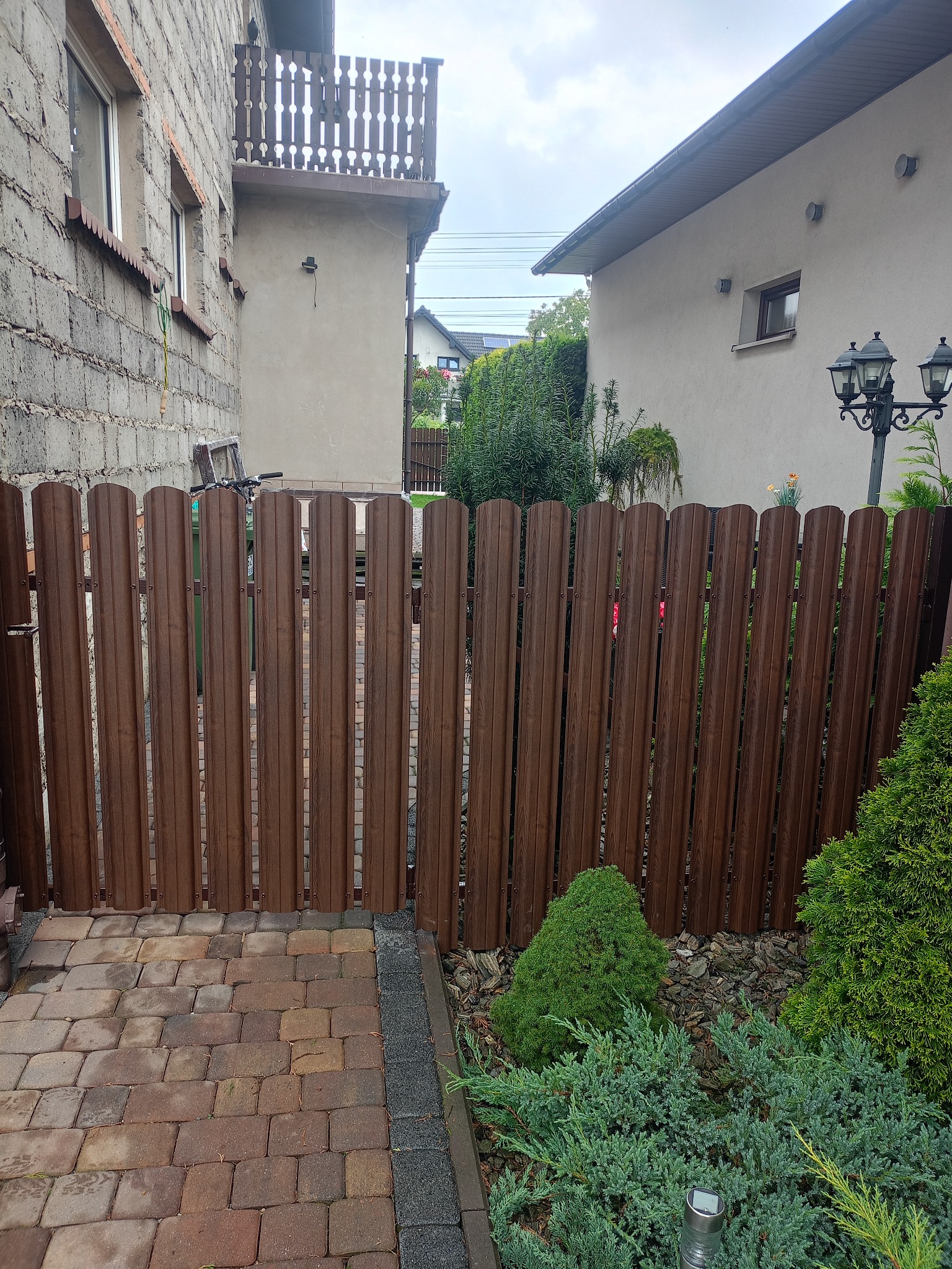 Brązowa furtka z pionowych paneli, wkomponowana w ogrodzenie, z kostką brukową i zielenią w tle, pomiędzy budynkami. Widok na podwórko.