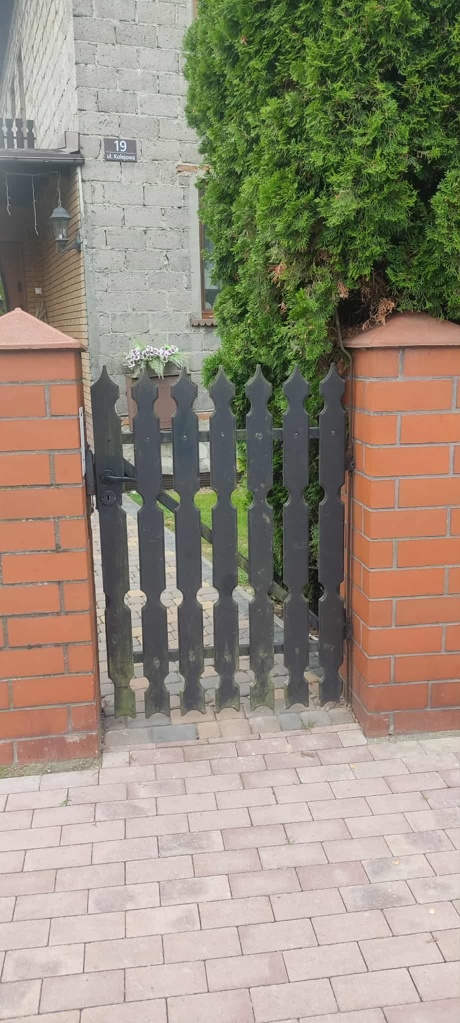 Ciemna furtka z dekoracyjnymi wycięciami, osadzona między ceglanymi słupkami, prowadząca na posesję z zielonym trawnikiem i roślinnością. Widoczny numer domu.