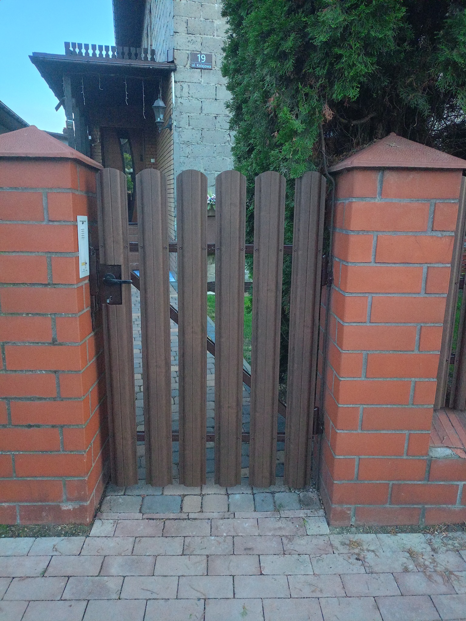 Brązowa furtka z pionowych paneli, osadzona między ceglanymi słupkami z daszkami, prowadząca do domu z numerem 19. Widok na chodnik z kostki.