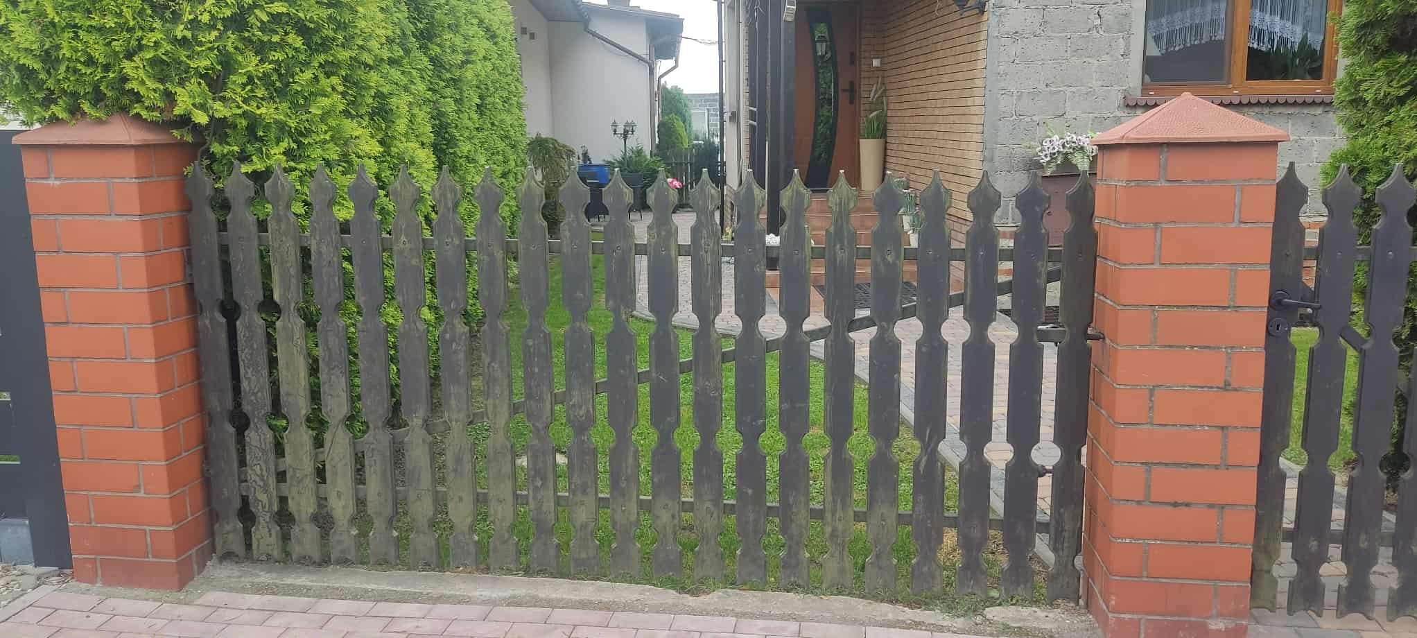 Ciemne, drewniane ogrodzenie z dekoracyjnymi wycięciami, wsparte na ceglanych słupkach z daszkami, na tle zieleni i domu z cegły. Widok z zewnątrz posesji.