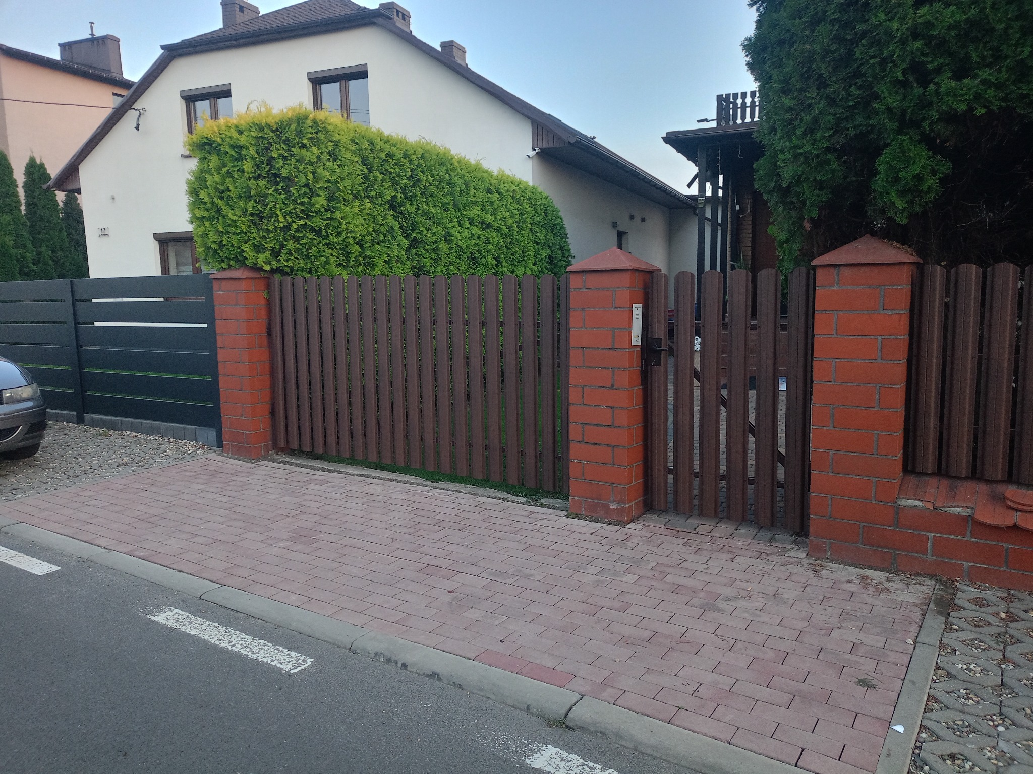 Nowoczesne ogrodzenie posesji z brązowych paneli i ceglanych słupków, furtka z tego samego materiału. Częściowo widoczny szary samochód i chodnik z kostki.