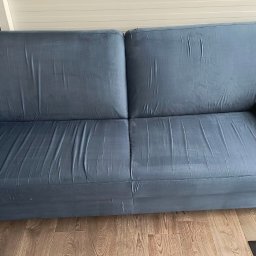 SKY CLEANING - Dwuosobowa, niebieska kanapa z poduszkami na drewnianej podłodze, widoczny fragment zasłony i wycieraczki.