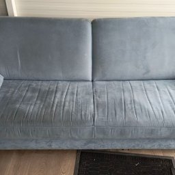 SKY CLEANING - Szara, dwuosobowa kanapa z poduszkami, widoczne ślady użytkowania na siedzisku, drewniane nóżki, obok drzwi balkonowe.