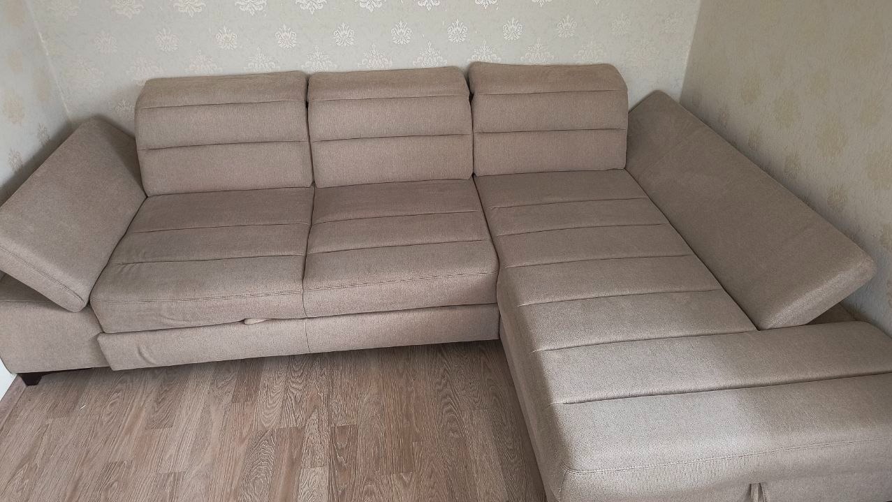 Beżowa, narożna kanapa z tapicerowanymi siedziskami i oparciami w salonie z drewnianą podłogą i wzorzystą tapetą.