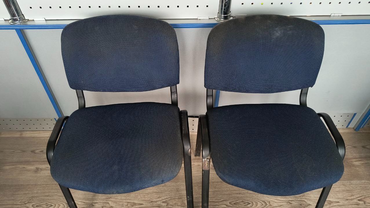 Dwa ciemnoniebieskie krzesła biurowe z czarnymi ramami, widoczne ślady użytkowania na siedziskach i oparciach, ustawione na drewnianej podłodze.