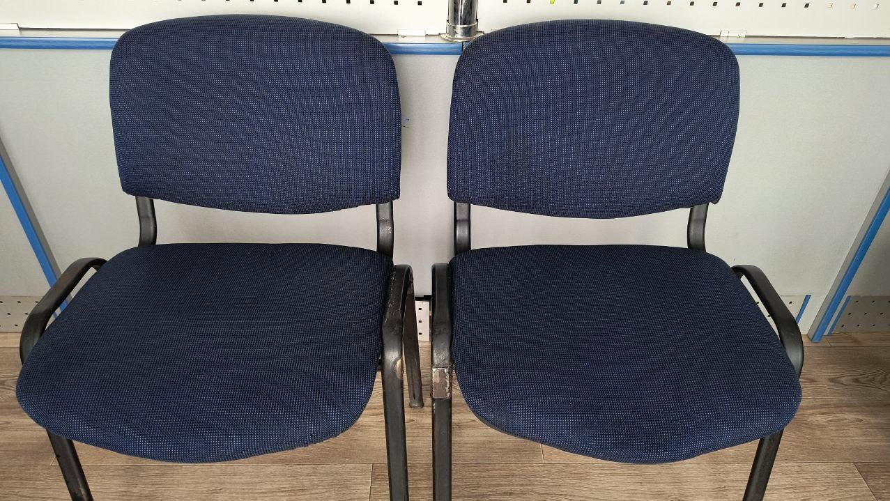Dwa identyczne krzesła biurowe z ciemnoniebieską tapicerką i czarnymi metalowymi ramami, ustawione obok siebie na podłodze imitującej drewno, przed białą ścianą z perforacjami i fragmentem...
