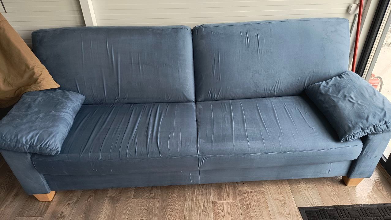 Dwuosobowa, niebieska kanapa z poduszkami na drewnianej podłodze, widoczny fragment zasłony i wycieraczki.