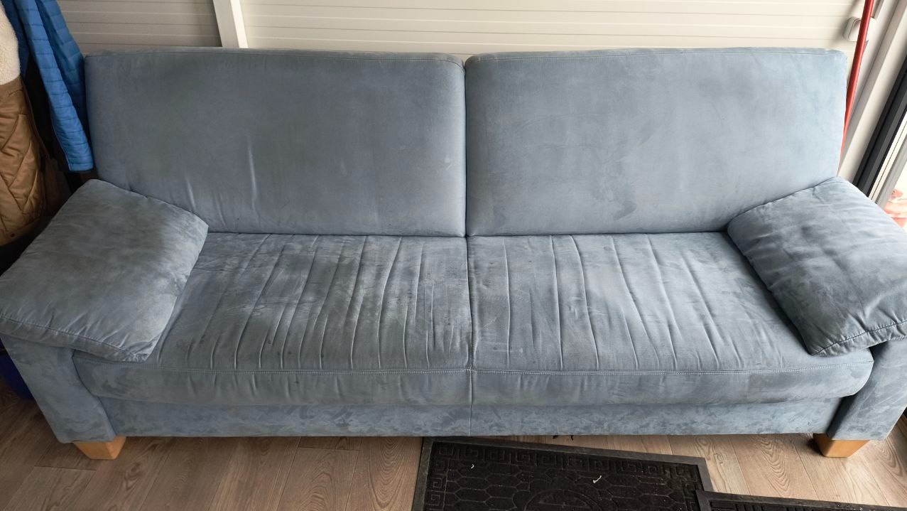 Szara, dwuosobowa kanapa z poduszkami, widoczne ślady użytkowania na siedzisku, drewniane nóżki, obok drzwi balkonowe.