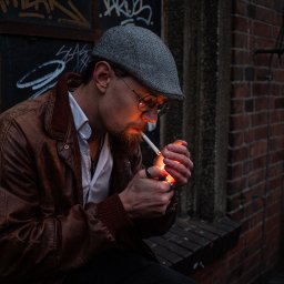 Dorian Durys - Mężczyzna z brodą w kaszkiecie i okularach zapala papierosa zapalniczką, na tle ceglanej ściany i graffiti.