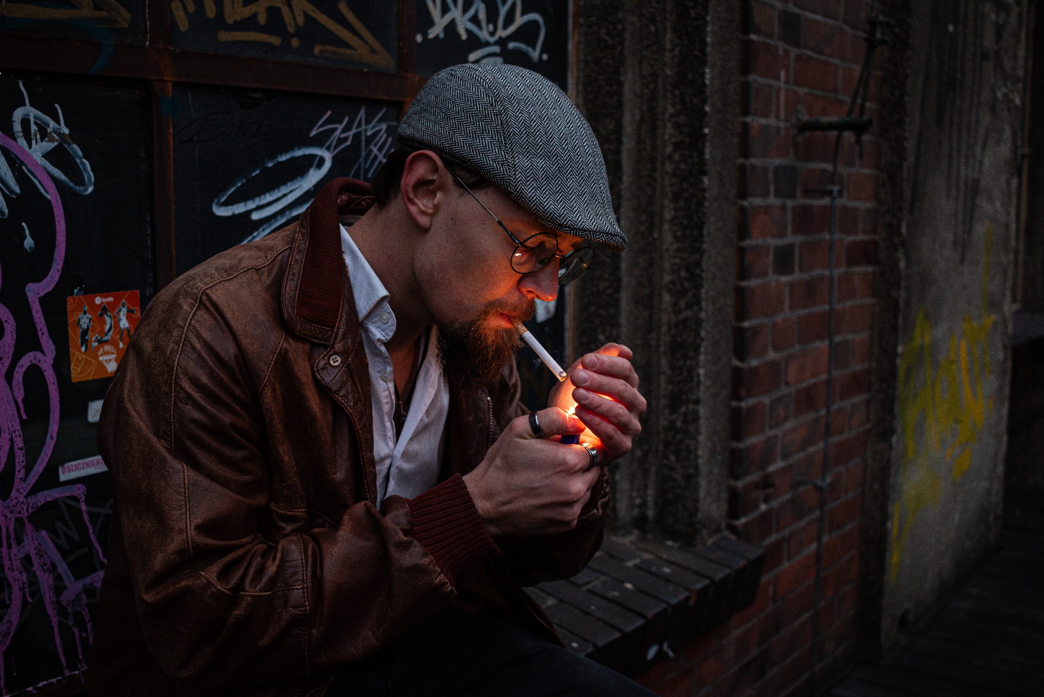 Mężczyzna z brodą w kaszkiecie i okularach zapala papierosa zapalniczką, na tle ceglanej ściany i graffiti.