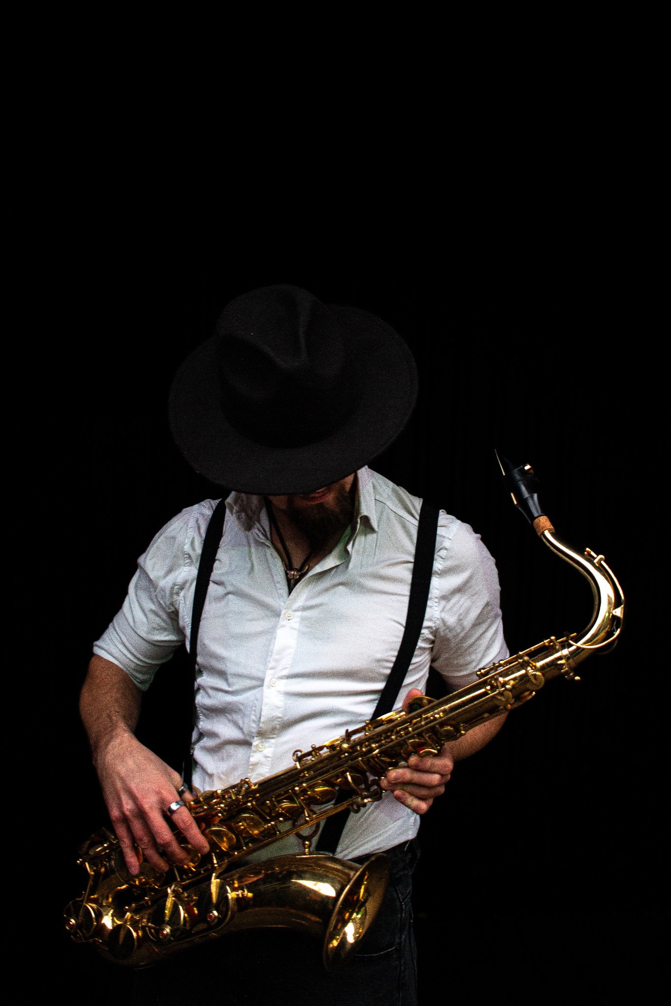 Saksofonista w białej koszuli i kapeluszu grający na saksofonie na czarnym tle, widoczne dłonie i fragment twarzy.