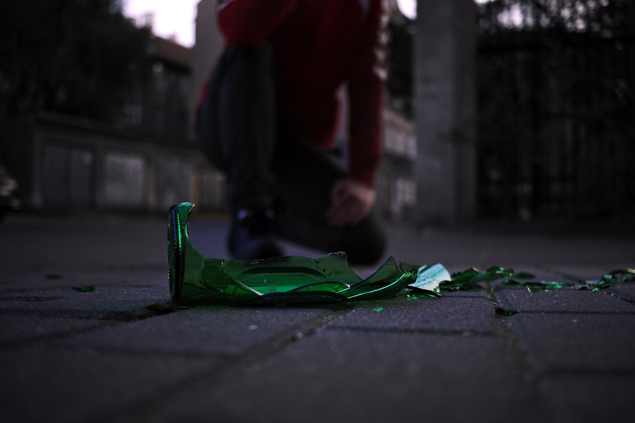 Rozbite, zielone szkło butelki leżące na brukowanej powierzchni; w tle niewyraźna postać klęcząca na chodniku.