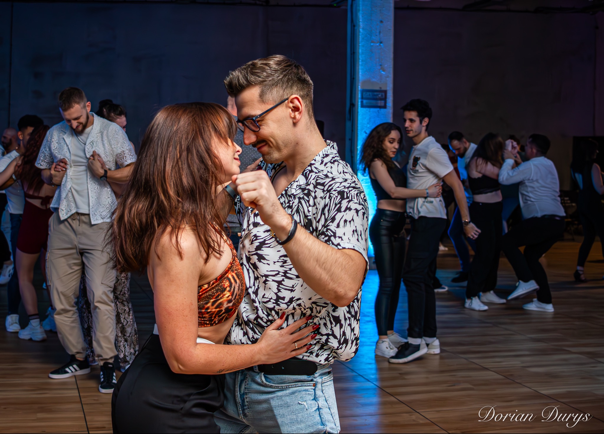 Para tańcząca salsę w pomieszczeniu z innymi parami w tle, mężczyzna w okularach i koszuli we wzory, kobieta w crop topie w panterkę.