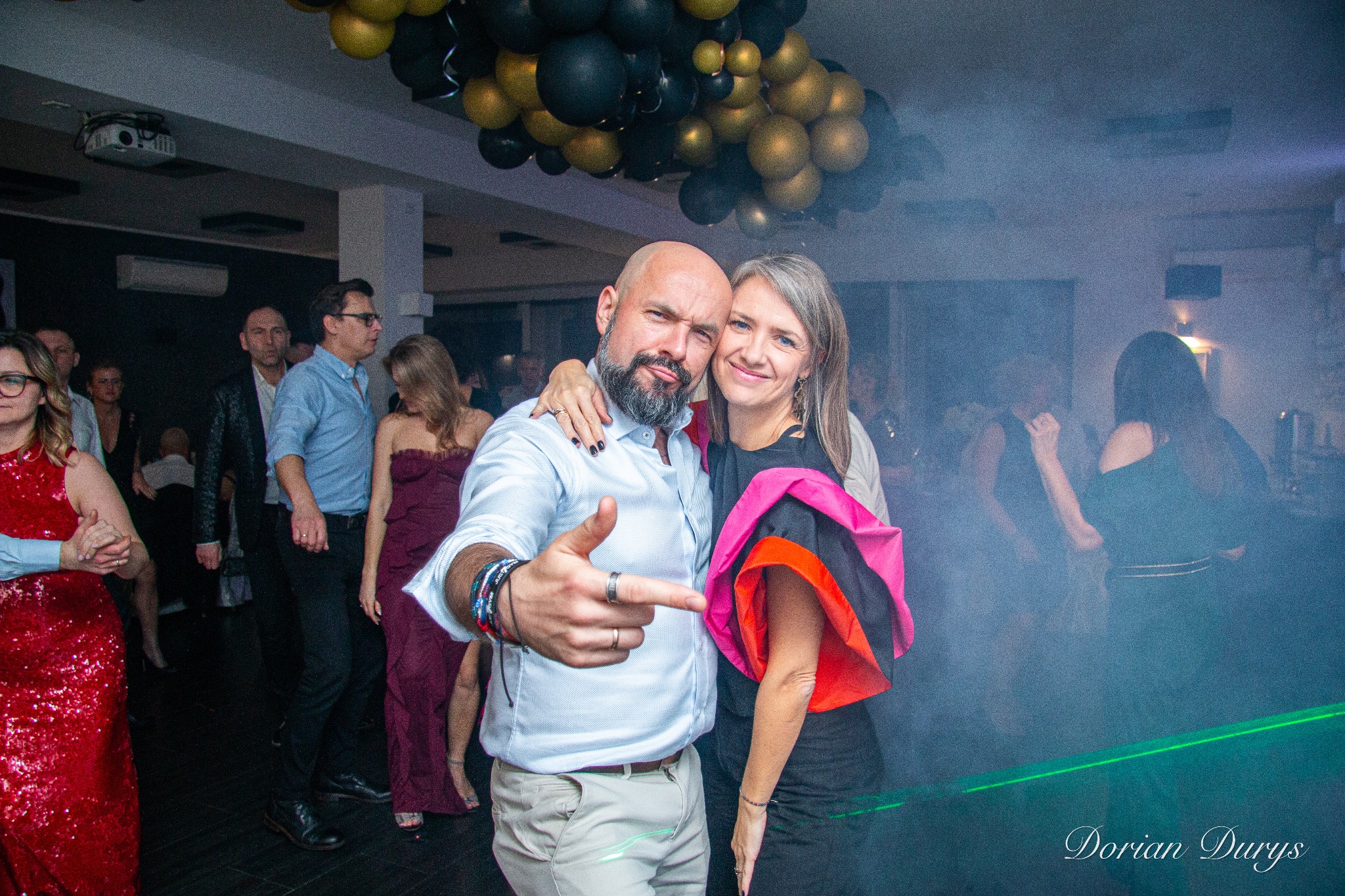 Para pozuje na tle sali balowej z dekoracjami z czarno-złotych balonów, mężczyzna z brodą wskazuje palcem w stronę obiektywu, w tle widoczni inni goście i efekty świetlne.