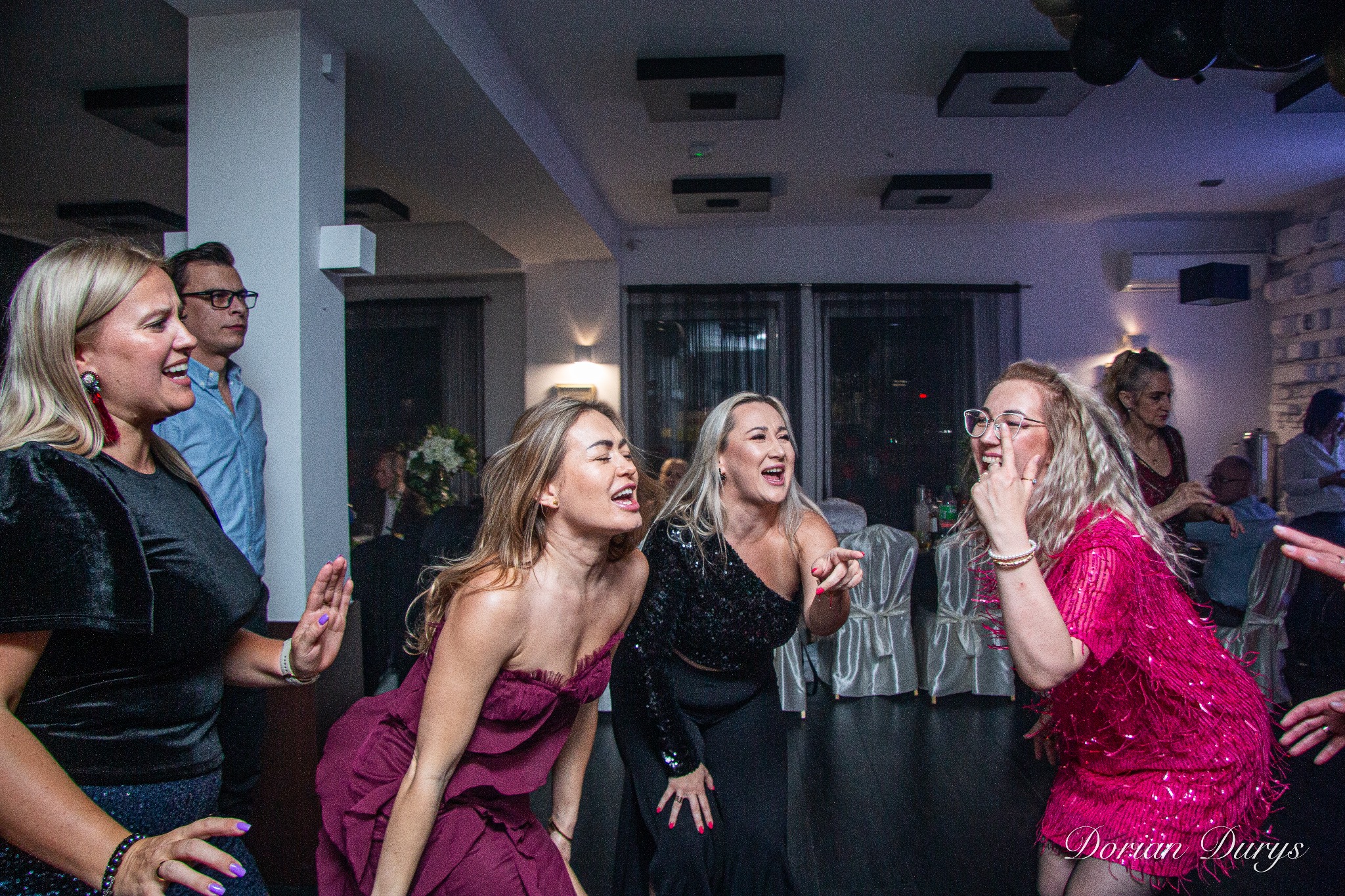 Rozbawione kobiety tańczą na imprezie, w tle widoczni inni goście i dekoracje sali.