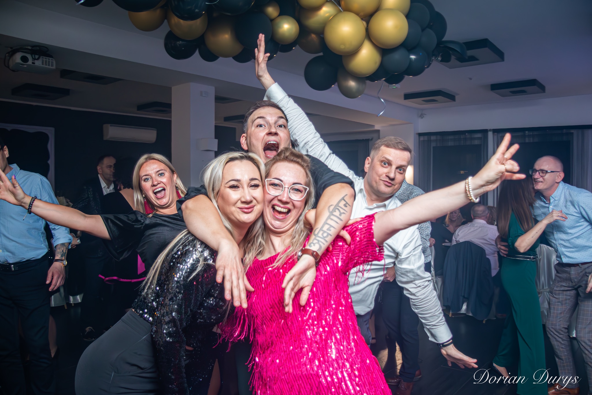 Grupa roześmianych osób pozuje do zdjęcia na tle sali z balonami, mężczyzna z tatuażem na ramieniu w centrum kadru.