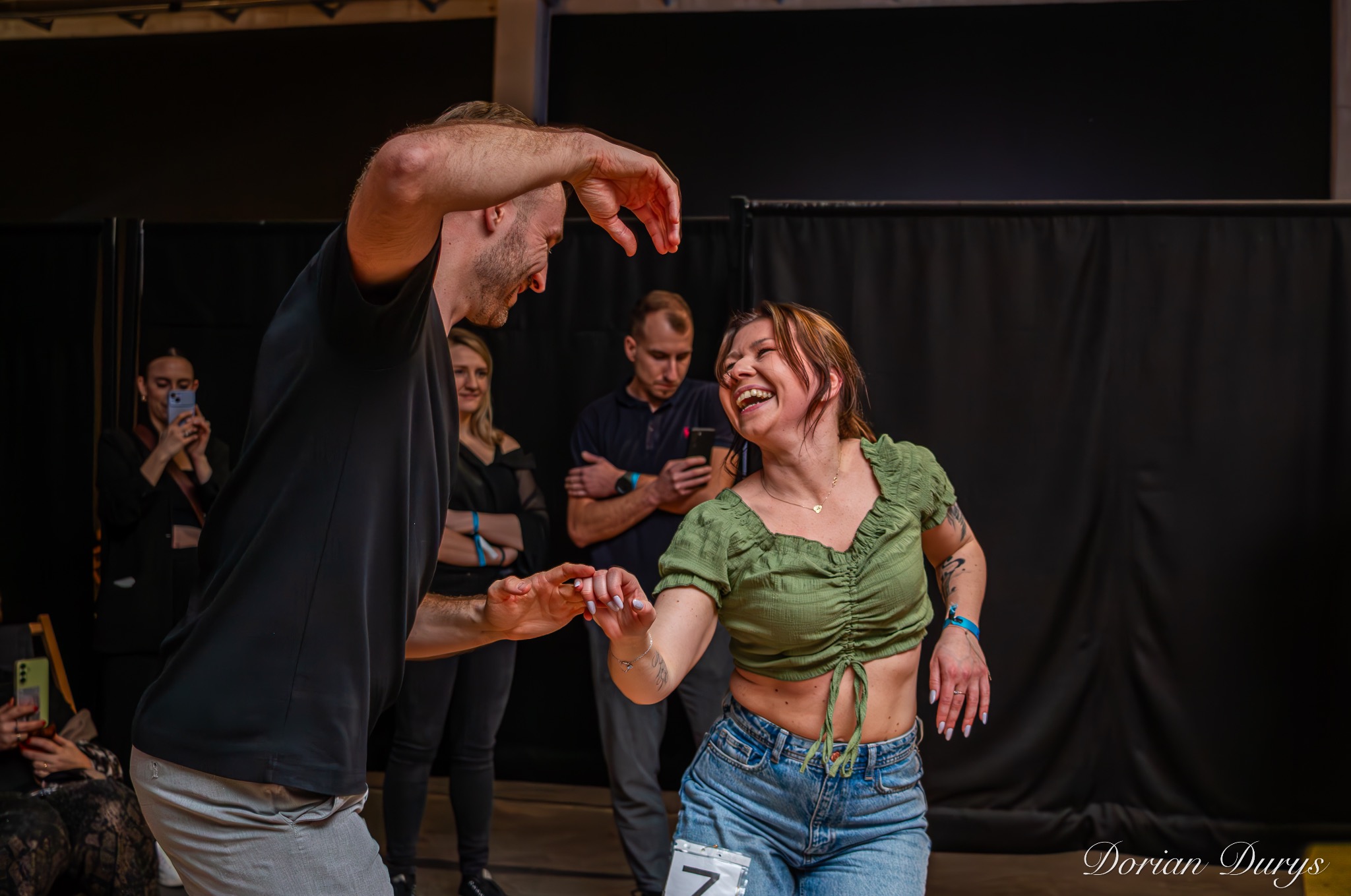 Para w Gdańsku tańczy swinga na tle czarnej kotary, kobieta w dżinsach i zielonej bluzce śmieje się, mężczyzna w czarnej koszulce prowadzi.